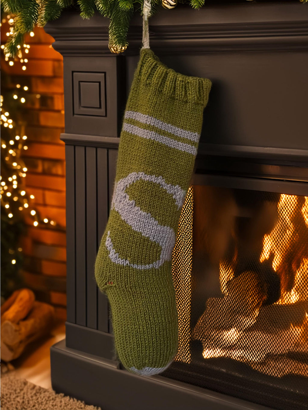 Army green hand-knit Christmas stocking with grey S letter by a lit fireplace