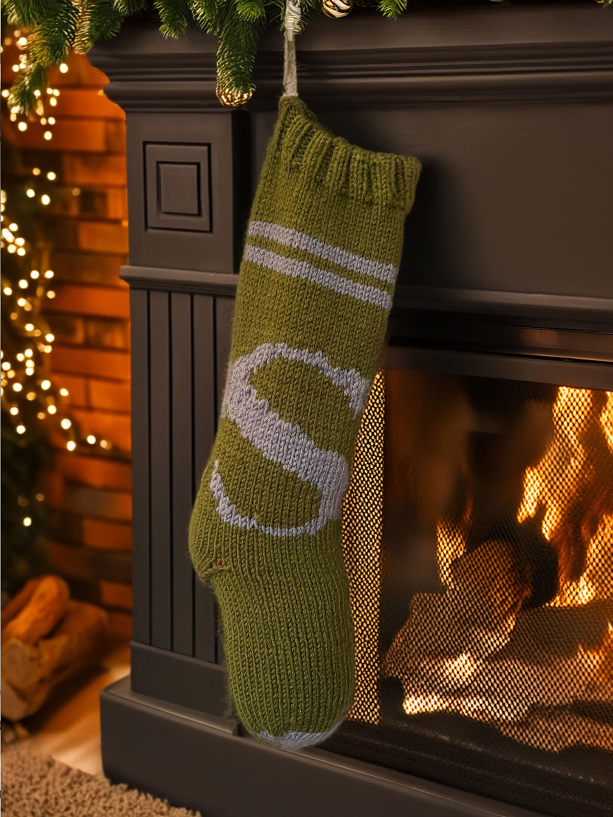 Army green hand-knit Christmas stocking with grey S letter by a lit fireplace