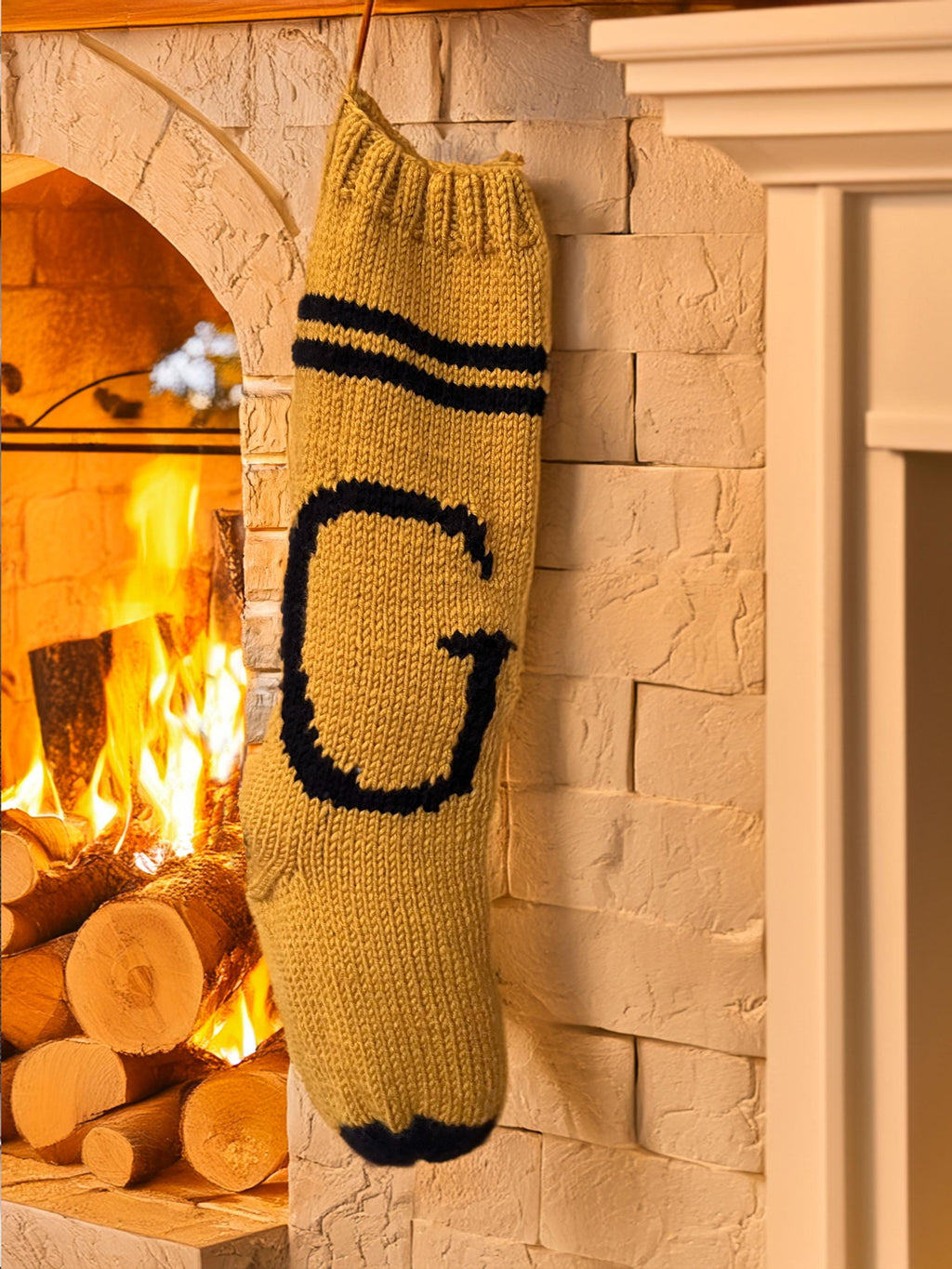 Gold hand-knit Christmas stocking with black G letter beside a stone fireplace — knit detail