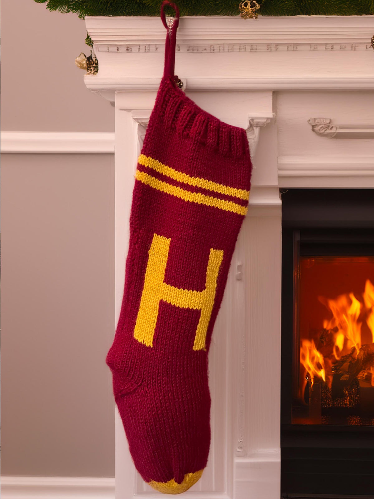 Burgundy hand-knit Christmas stocking with gold H letter and stripes, hanging by the fire