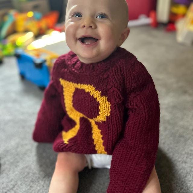 Smiling baby in burgundy monogram sweater with gold R letter — hand-knit baby jumper