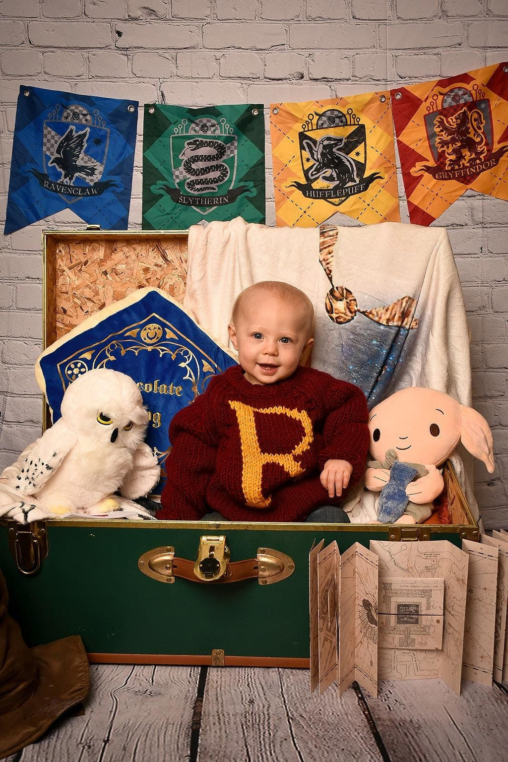 Baby wearing burgundy monogram sweater with gold R letter, sitting in a vintage trunk